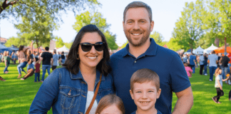 A smiling family of four poses on vibrant green grass at a busy outdoor community event surrounded by other attendees and colorful vendor tents under a clear blue sky